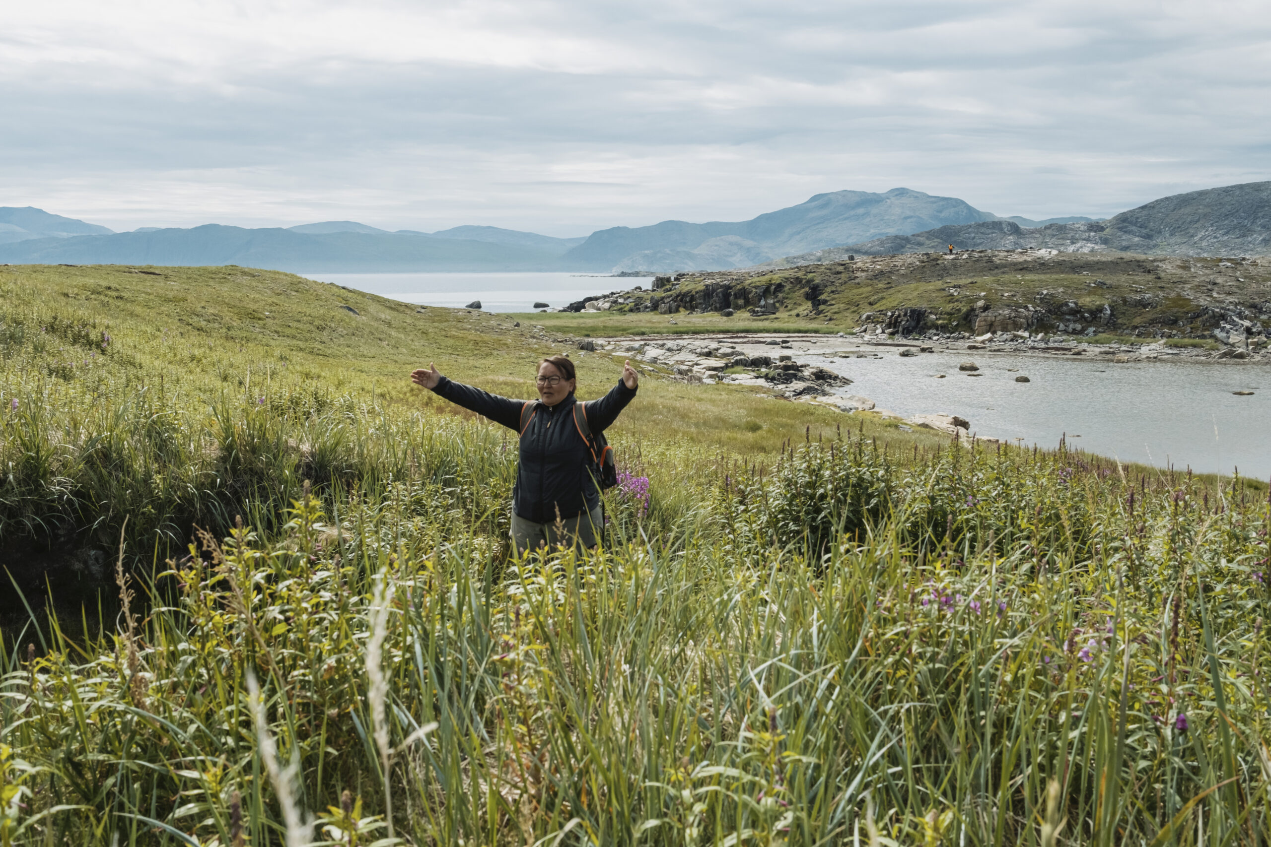 Torngat Mountains National Park: A woman raises her arms showing the edges of a depression in tall grass with Torngat Mountains in the background