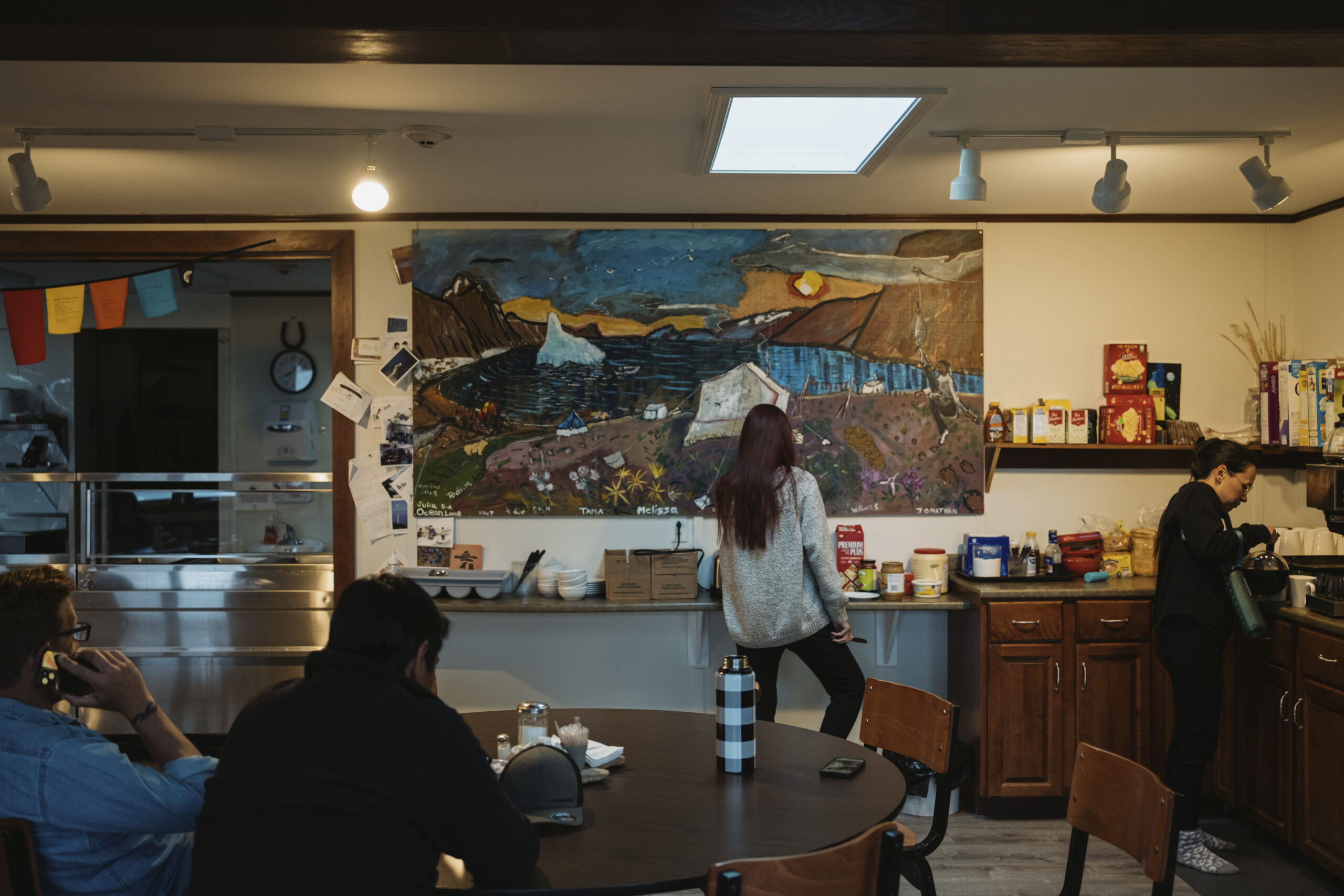 People sit around a table in the foreground as a woman pours coffee and another woman stands at a counter with a painting and decorations in front of her.