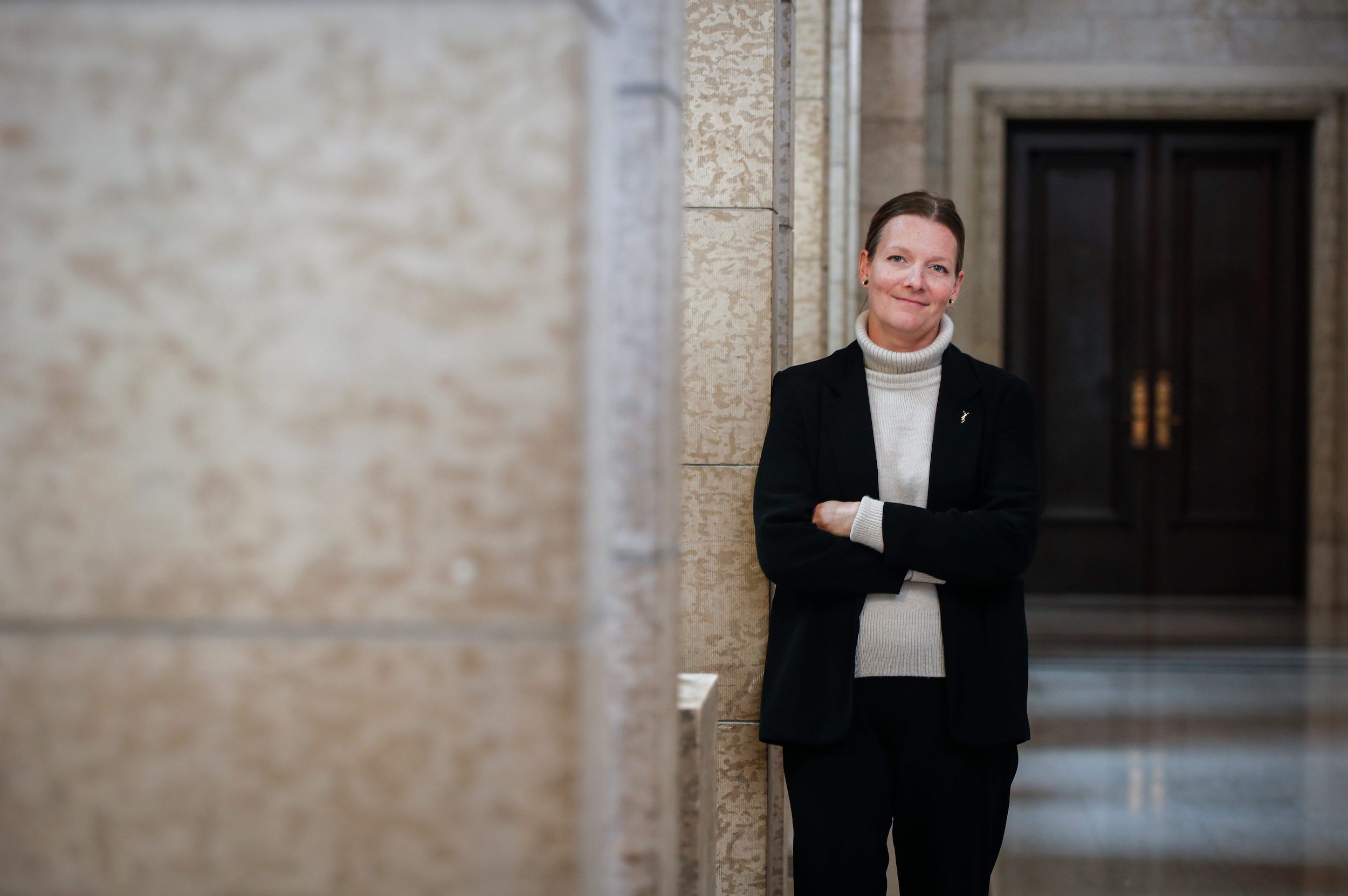 Tracy Schmidt wears a black blazer and beige turtleneck standing in the halls of the Manitoba legislative building