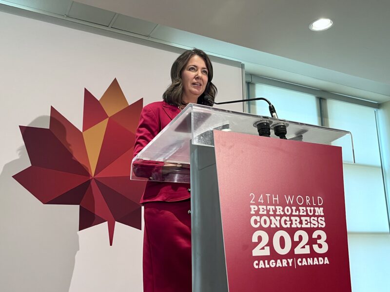 Alberta Premier Danielle Smith, standing at a lectern, has said the renewables pause was spurred by requests from the grid manager and regulator