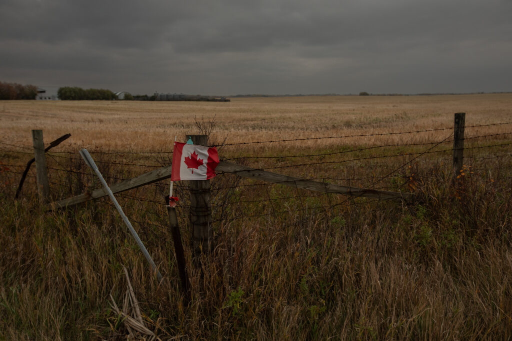 Canadian flag on a post in front of a barbed-wire fence enclosing a farm-field in Alberta