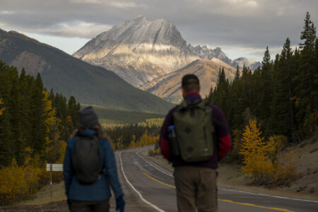 Hikers carrying backpacks walk alongside a highway in Kananaskis with a large mountain peak looming ahead