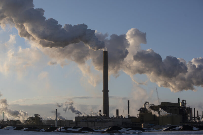 Large plumes rise into blue skies above a stack in Alberta's oilsands, with equipment in the foreground