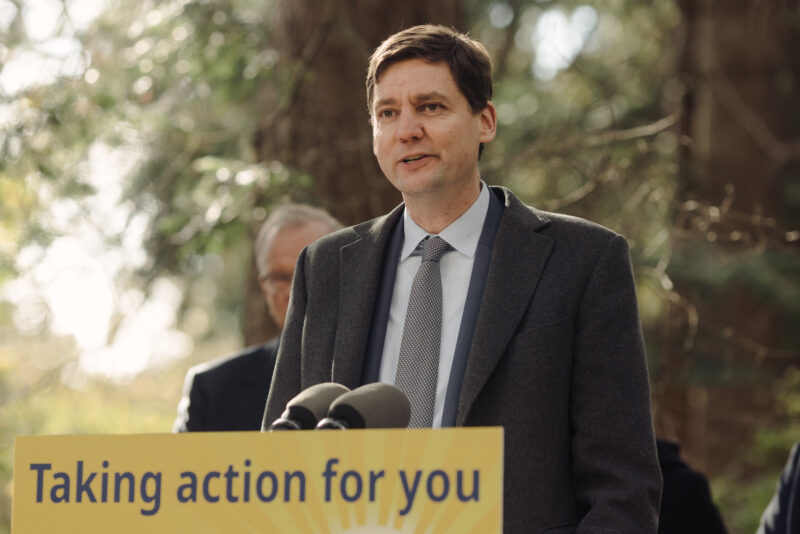 B.C. Premier David Eby speaking at a podium with trees in the background