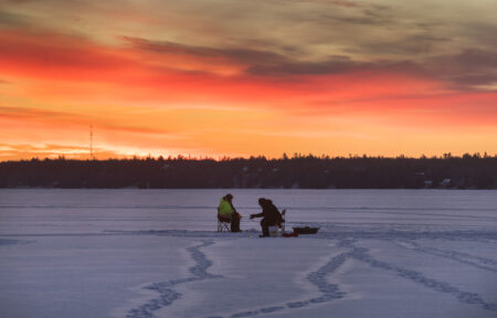 Two anglers sit on a frozen lake over an ice-fishing hole under an orange and red sunrise