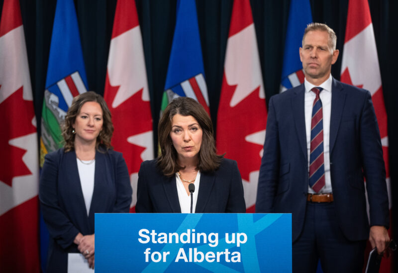 Alberta Premier Danielle Smith stands at a lectern, flanked by ministers Rebecca Schultz and Nathan Neudorf