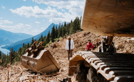 Eddie Petryshen and Sarah Cox stand amid a clear-cut with construction equipment in the foreground in B.C.'s inland temperate rainforest, overlooking the Revelstoke reservoir.