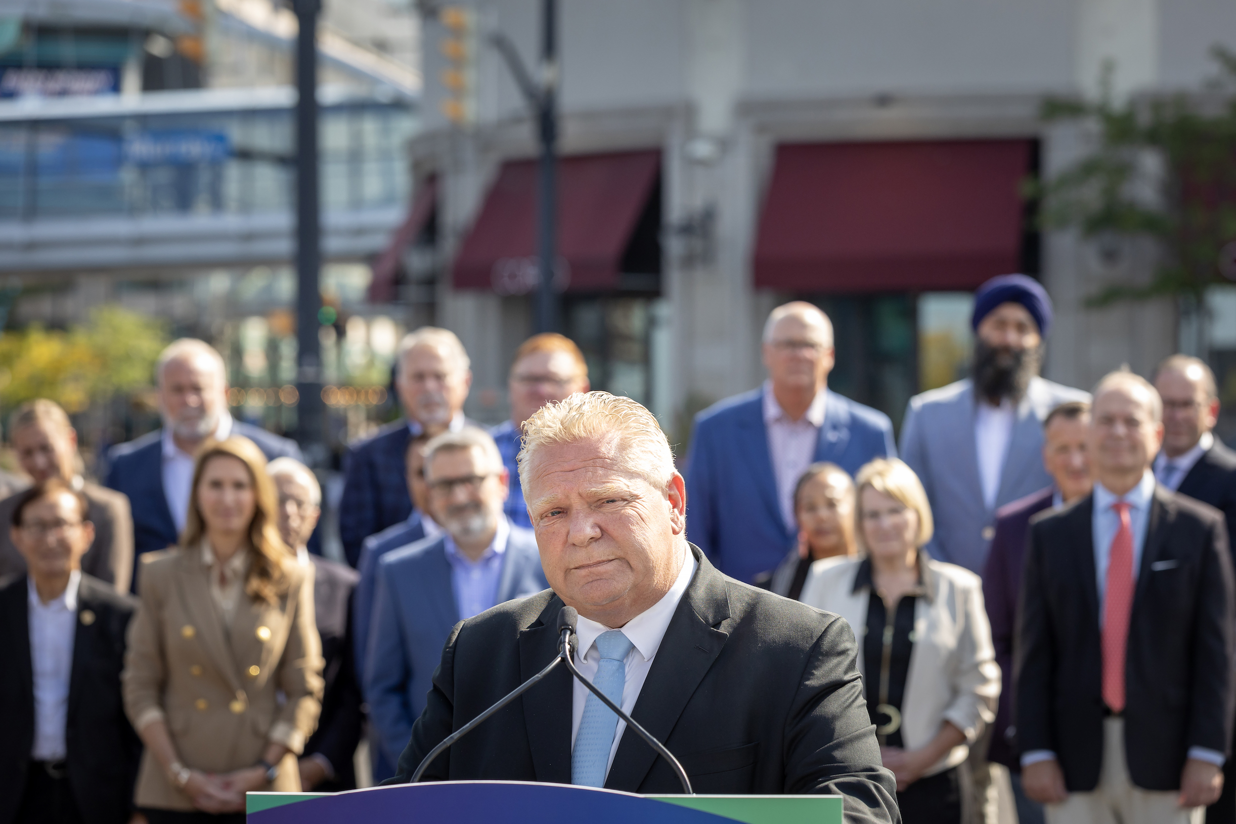 Doug Ford with a solemn expression at a microphone, with his cabinet standing behind him