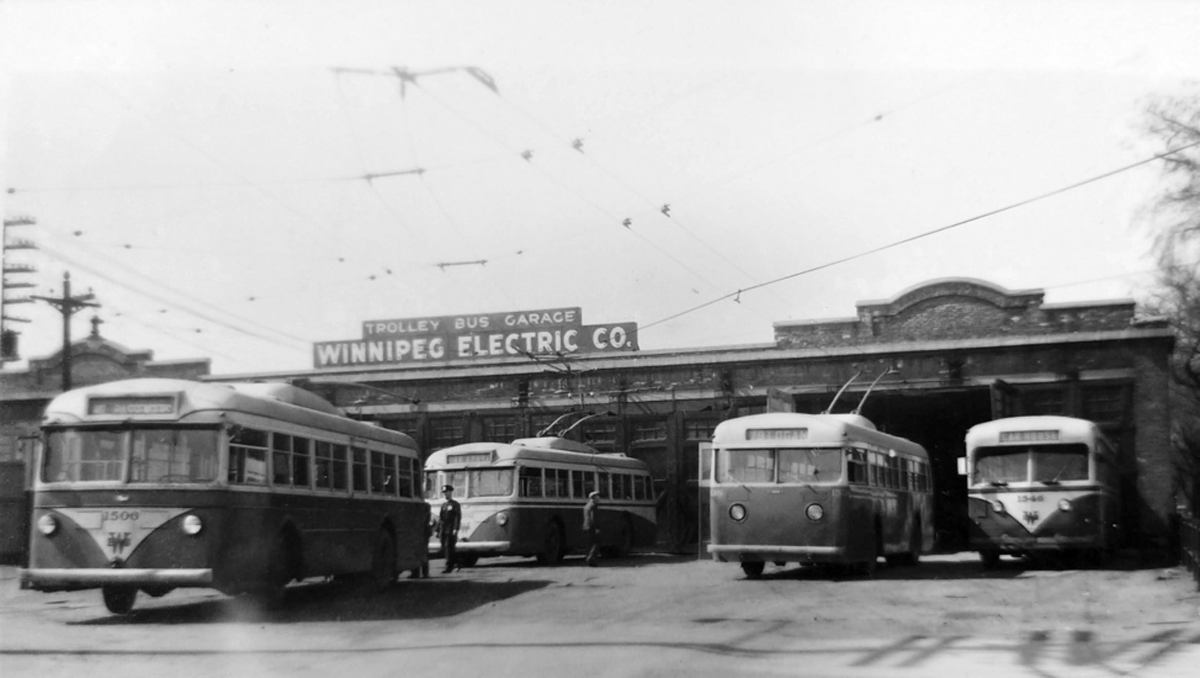 A black and white archival image shows four electric trolley buses at a Winnipeg Electric Company garage on Assiniboine Avenue