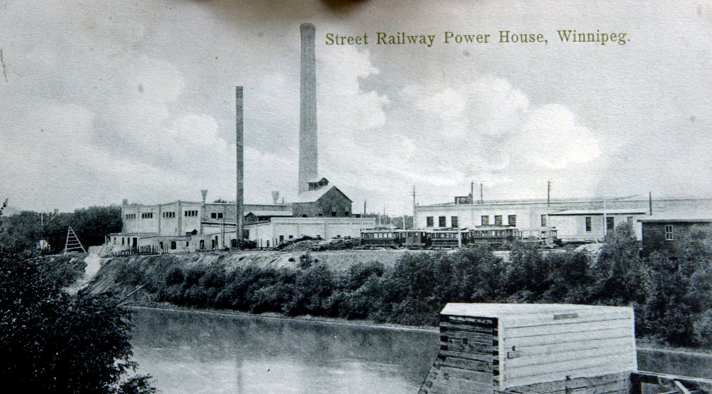 A black and white card labeled 'Street Railway Power House, Winnipeg' depicts a steam coal fired power generating station and streetcar garage, with a few parked streetcars, from across the Assiniboine River