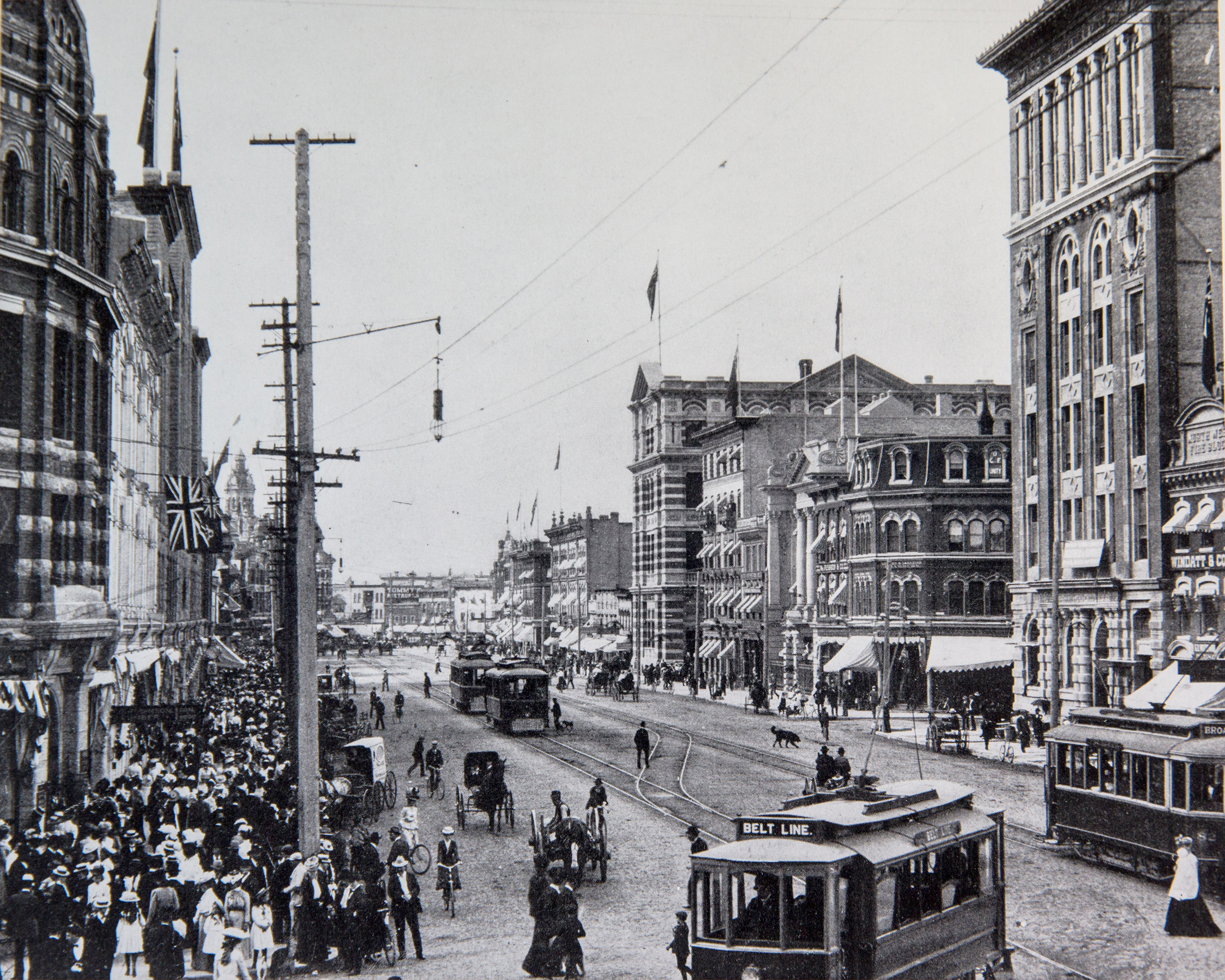 A streetcar with a sign reading "Belt Line" turns off of Main Street in Winnipeg. Behind it, other streetcars and horse-drawn carriages use the road while a large crowd of people gather on the sidewalks in 1905