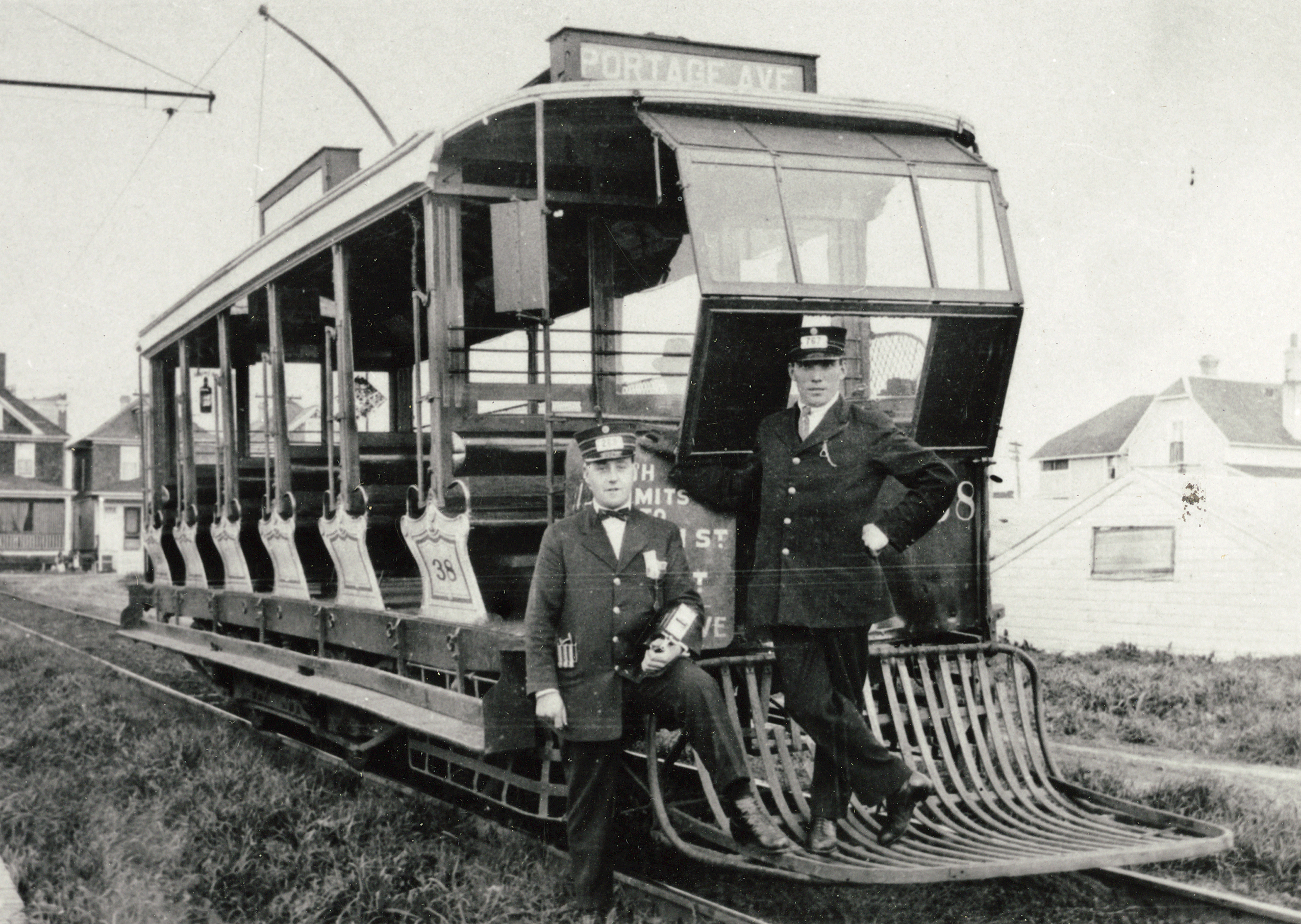 Two men in suits stand on a rack extending from the front of an open carriage streetcar with a sign reading "Portage Ave" in Winnipeg
