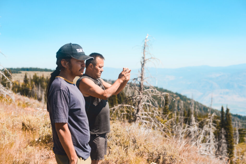 Tim Lezard and Weston Roberds stand atop a mountain monitoring a wildfire outside the frame. Dry grasses and forests stretch across the hillsides around them