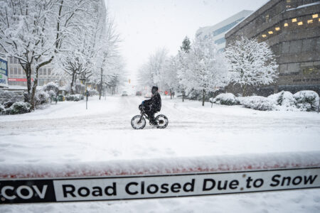 Vancouver street covered in snow, with a sign that reads 'road closed due to snow' in the foreground. The trees, ground, and sky are white. In the centre, a person rides their bike down the middle of the empty snow-covered road during the January 2024 first snow in Vancouver.