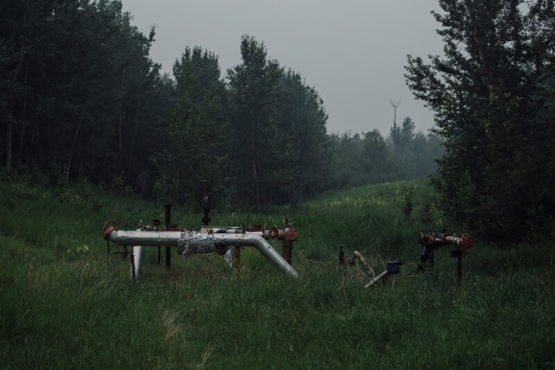Deteriorating oil and gas infrastructure in Alberta in a clearing of green grass surrounded by low trees and smoky skies