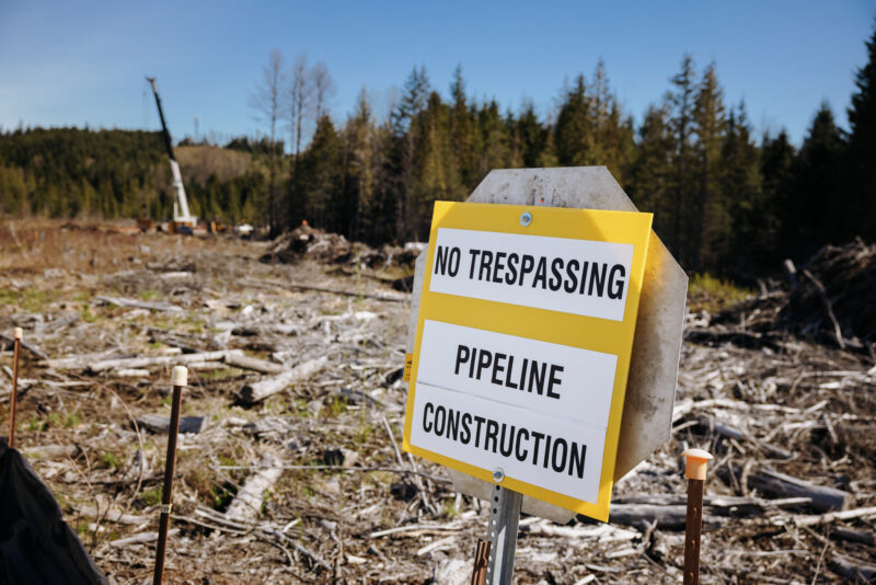 A yellow and white sign that says "No tresspassing, pipeline construction" in front of a cleared area of a forest with a crane in the background.