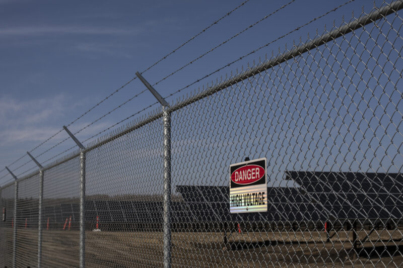 A solar installation sits behind a chain link fence topped with barbed wire, with a sign that says danger, keep out.