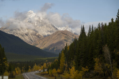 A road runs through a mountain valley in Kananaskis, Alberta, with treed slopes on either side and a cloud-shrouded mountain the background.