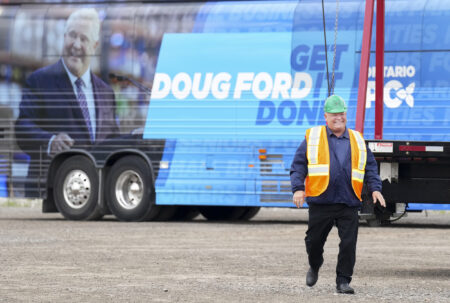 Doug Ford in a construction vest and hat walks in front of his campaign bus which says "Get it Done"