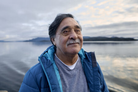 In Namu, B.C., on the central coast, Hereditary Chief Harvey Humchitt stands in the centre against a calm beach, and the glassy water reflects the overcast sky. Mountainous islands are visible on the horizon. Humchitt has dark hair with a bit of grey and a greying mustache. He wears a blue rain jacket and looks off camera to the right. Hhe has a soft expression and his eyes look deep and reflective