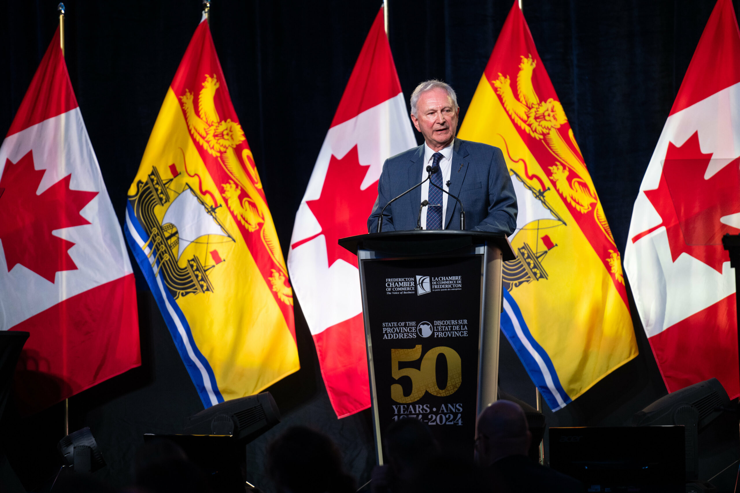 Man in a blue suit stands at a podium in front of Canadian and New Brunswick flags.