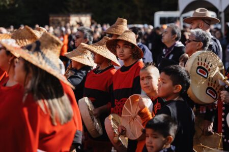 A crowd of people gathered to witness the rematriation of the pole, all wearing Nisga'a regalia, some holding drums. One person looks at the camera.