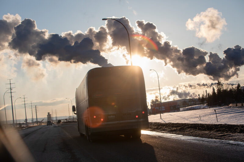 A bus emits smoke travelling along a highway under a blue sky and sunshine