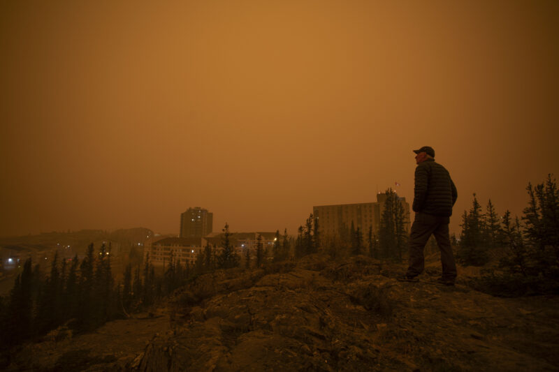 A man stands on a rocky outcrop looking out at the skyline of downtown Yellowknife under a dark orange sky, caused by wildfires