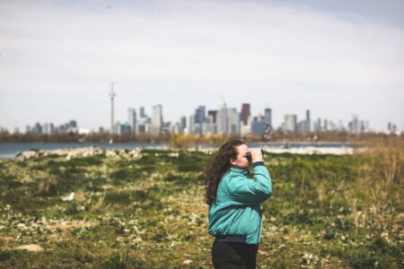 Emma McIntosh, wearing a teal jacket, peers through binoculars over a grassy landscape. The Toronto skyline is on the horizon.
