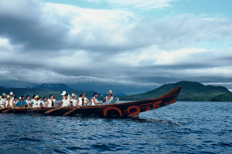 Dozens of people sit and paddle in a large Haida canoe on a blue ocean, with rolling green hills and cloudy skies.