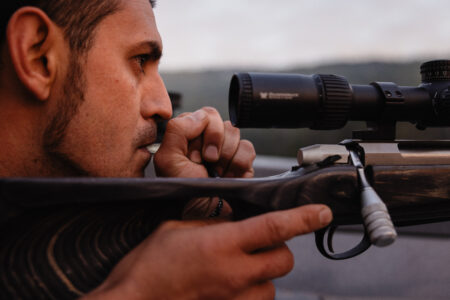 Lake Babine member Willie Bertacco holds an elk call device to his mouth. The camera is intimately close, and his face is in perfect profile as he gazes intently off camera to the right. His right hand holds a rifle at the ready. The scope is in front of his face. It's a tense, quiet moment as he prepares for his shot. The early morning sky is soft and pale grey and out of focus in the background