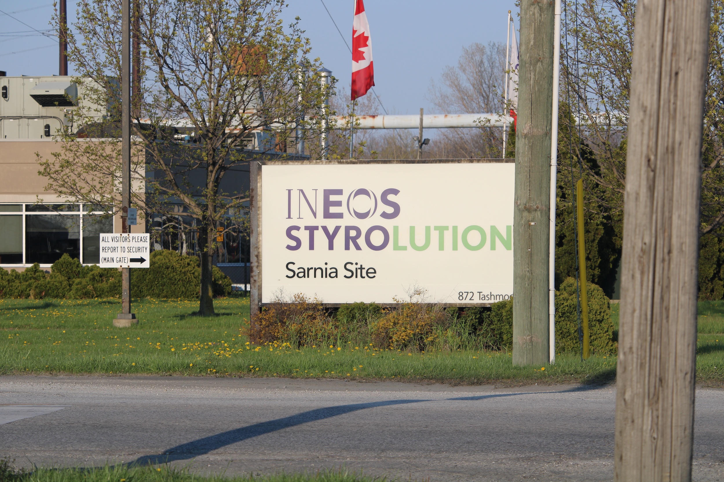 A large sign in front of office buildings reads "INEOS Styrolution Sarnia Site. A Canadian flag flies behind.
