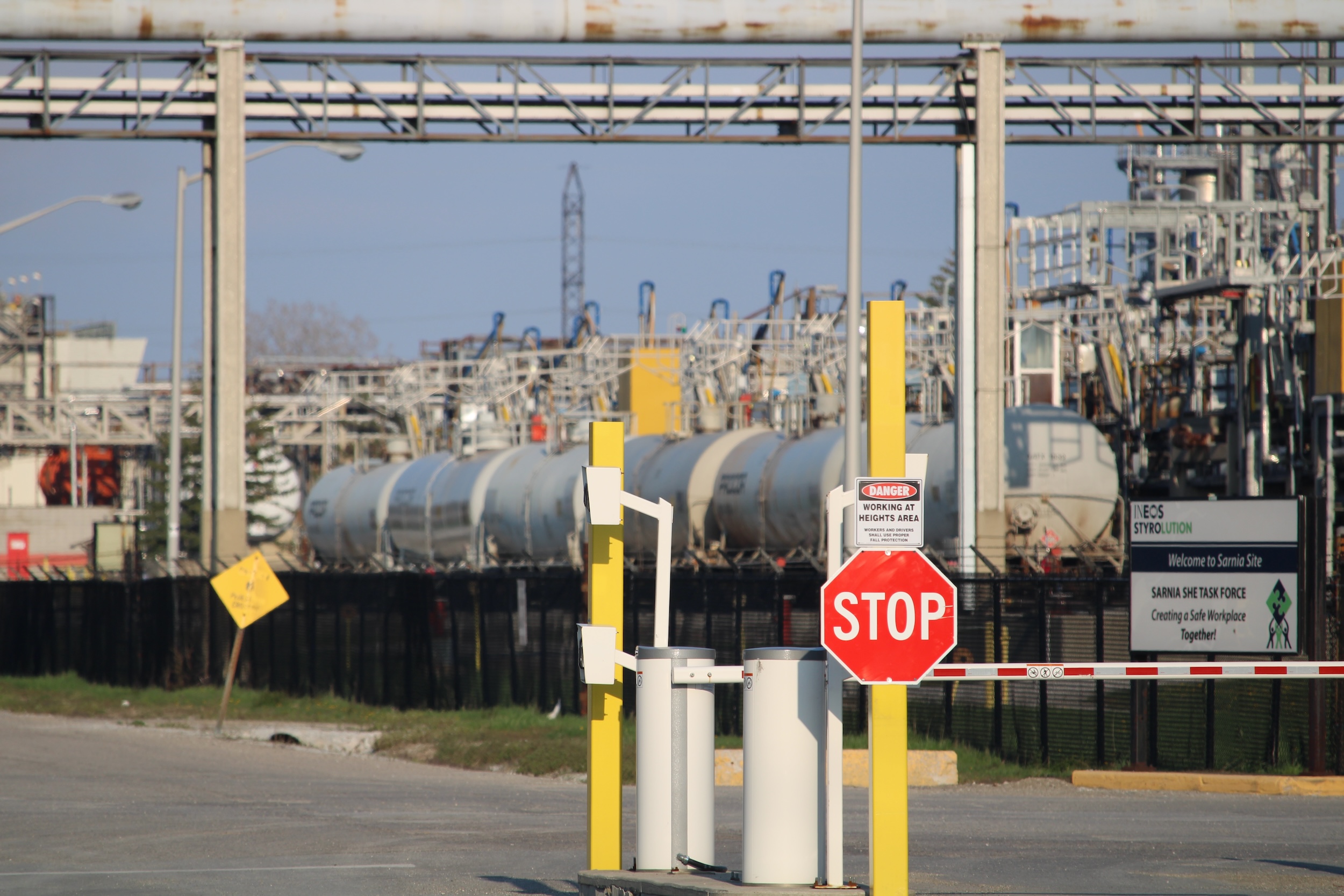 A stop sign and security gate at the entrance of an industrial facility. An array of industrial infrastructure fill the background.