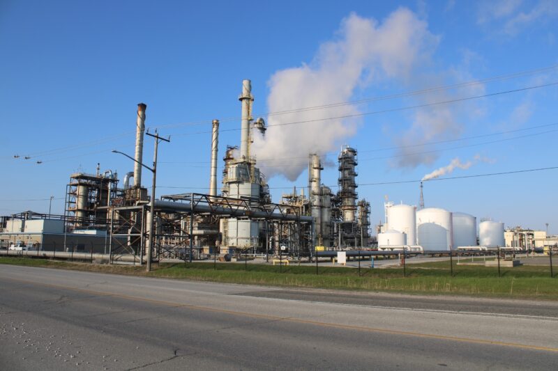 In Chemical Valley, louds of exhaust from an industrial factory punctuate blue skies.