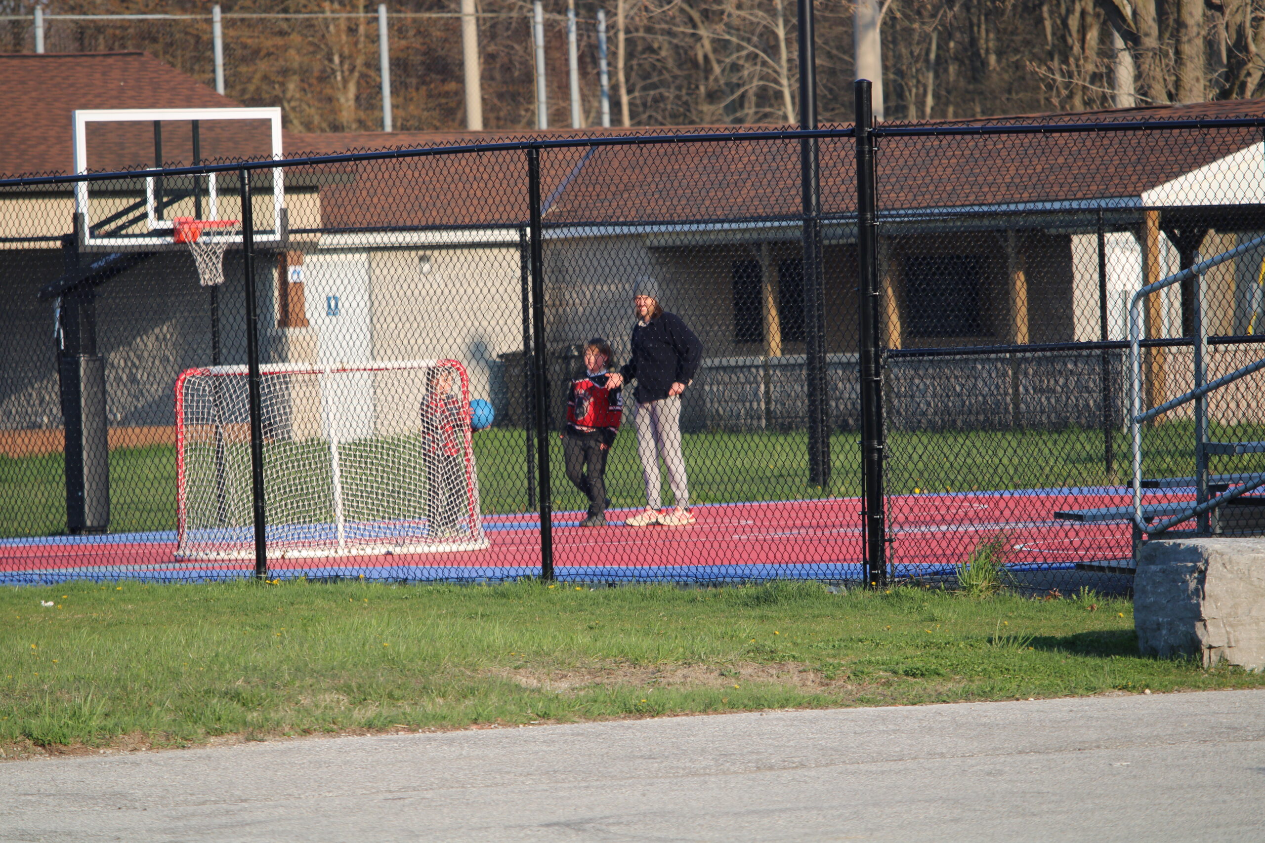 An adult and two children play with a basketball on an outdoor sports court, enclosed with high wire fences.