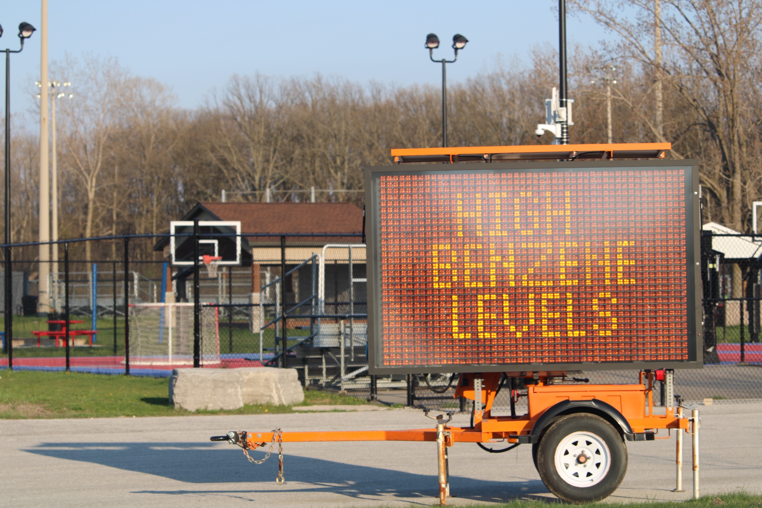 An electronic sign on an wheeled orange chassis reads, "HIGH BENZENE LEVELS." There are outdoor recreation fields and sport courts behind.