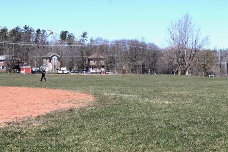 A man flies a kite at the edge of a baseball diamond while a group of children practice on another diamon in the background