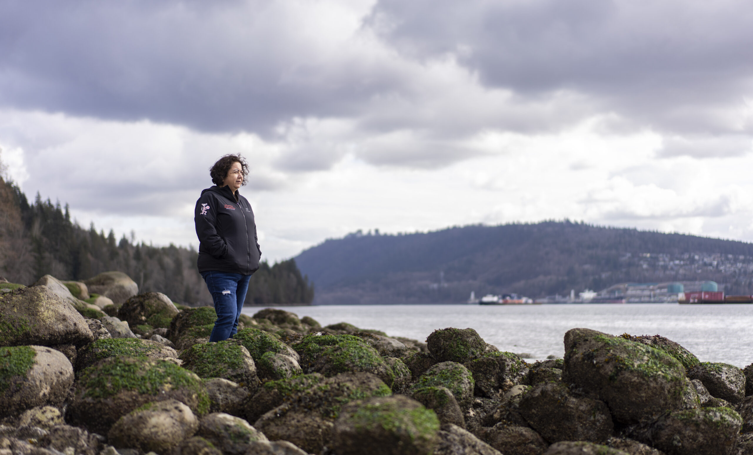 A portrait of Deanna George seated on a log on a rocky beach on the inlet, with mountains and part of the Westridge Marine Terminal visible on the opposite shoreA portrait of Deanna George standing on a rocky beach on the inlet, with mountains and part of the Westridge Marine Terminal visible on the opposite shore