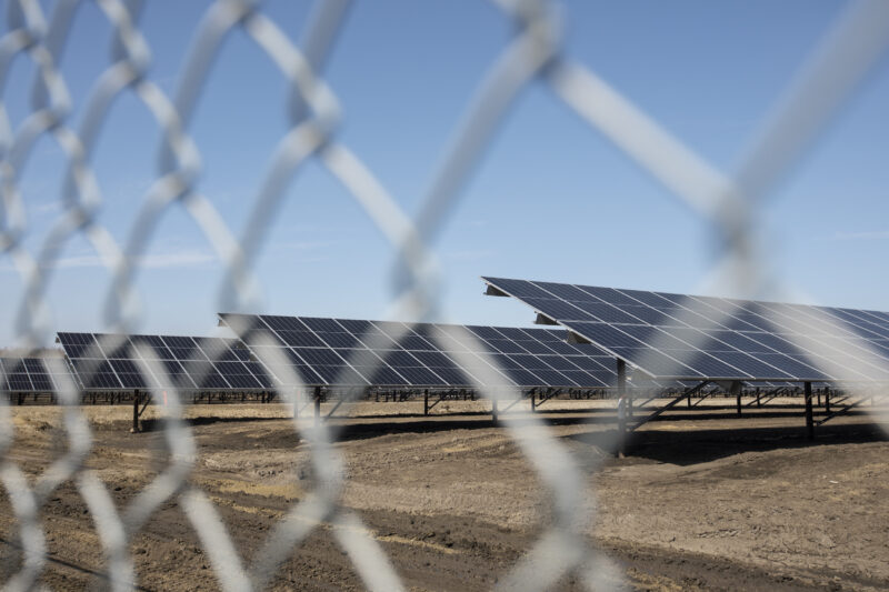 A large array of solar panels on brown dirt with blue skies, photographed through a wire fence.