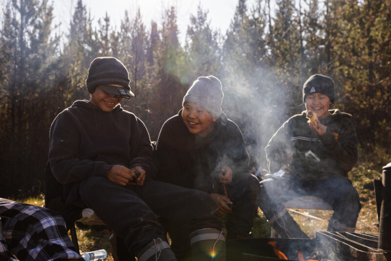 Three Lake Babine youth sit in a row on a log smiling and laughing, dressed warmly in tuques and sweaters. Sun comes down through the trees behind them, and campfire smoke rises in front of them