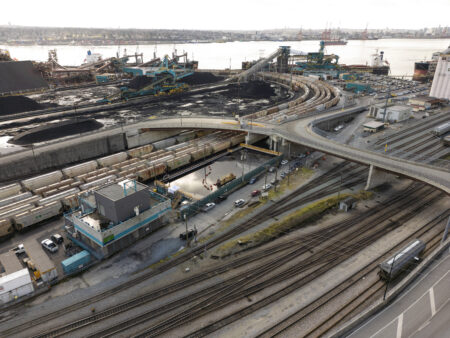 A photo of Neptune Terminals coal loading facility, with piles of coal and trains in teh foreground, and Burrard Inlet and Vancouver visible in the background