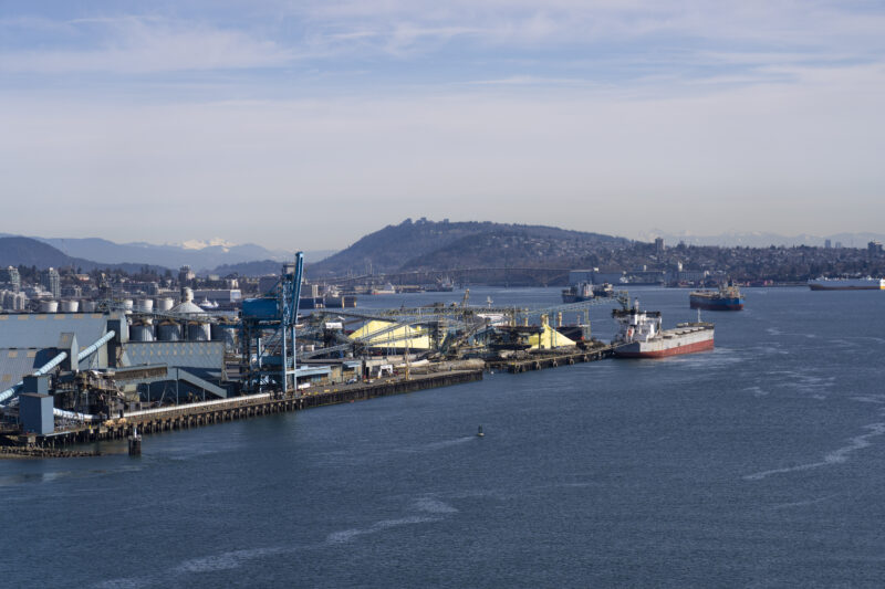 A photo of Burrard Inlet with boats traveling through the inlet, a ship is seen at Vancouver Wharves, with cities and mountains in the distance