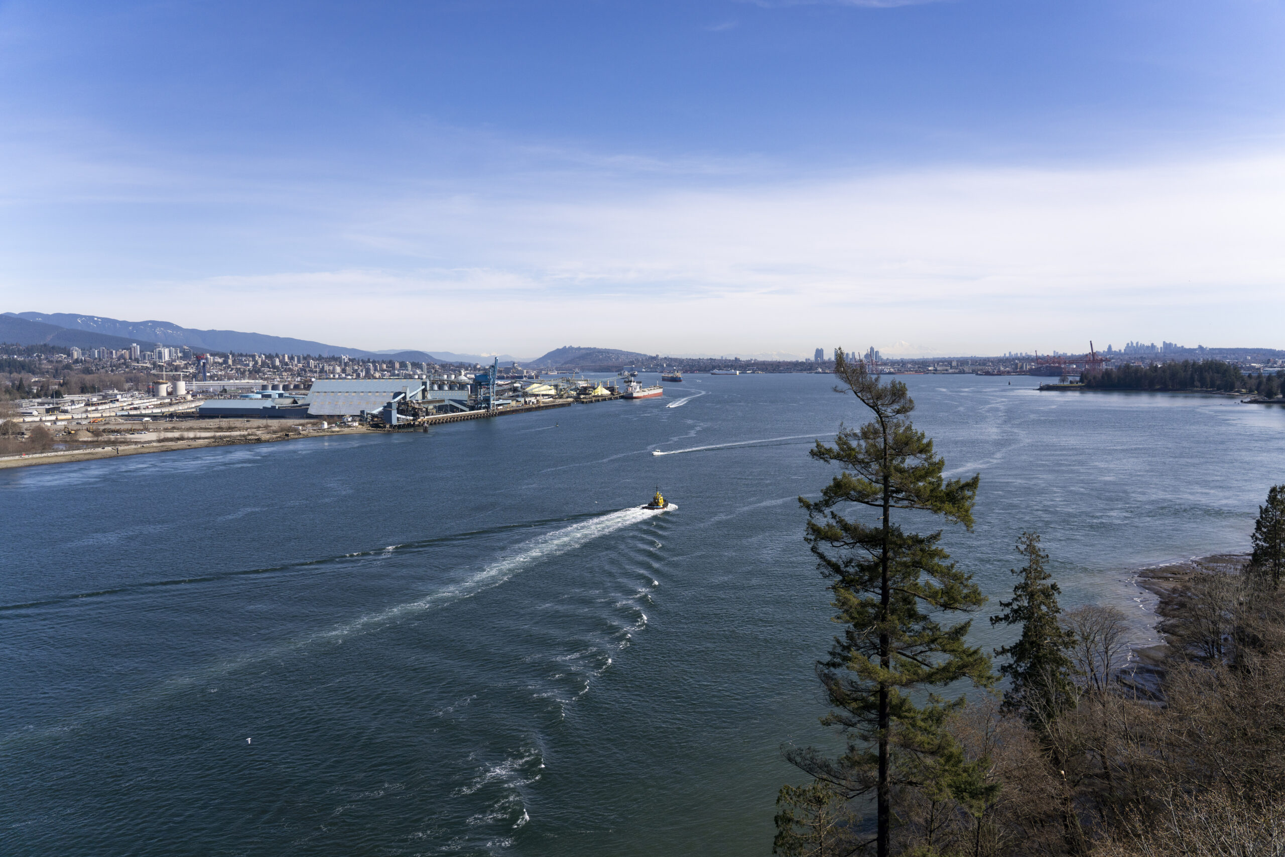 A photo of Burrard Inlet with boats traveling through the inlet, a ship is seen at Vancouver Wharves and the towers are seen in the background on both shores of the inlet