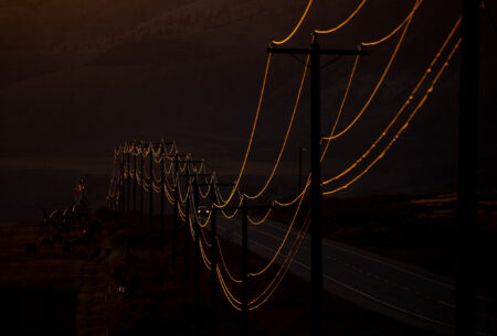 Amidst darkness, power lines gleam as they reflect the setting sun as a single car travels along a dark highway