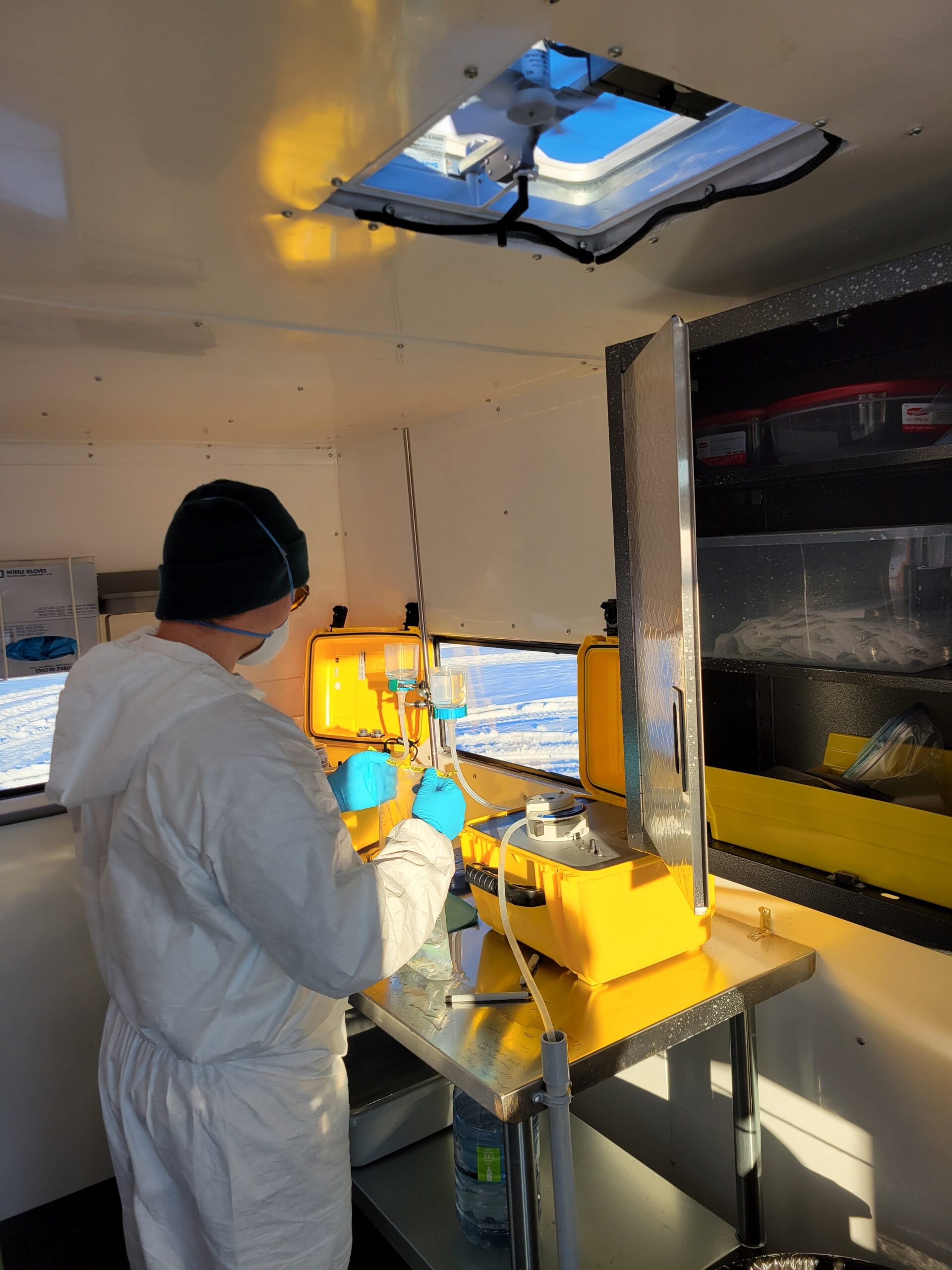 Riding Mountain National Park: A Parks Canada staff member in a white hazmat suit in a lab wears gloves and a mask n a heated shelter on a cold Manitoba day for the winter eDNA monitoring program, with a snowy scene visible outside a small window