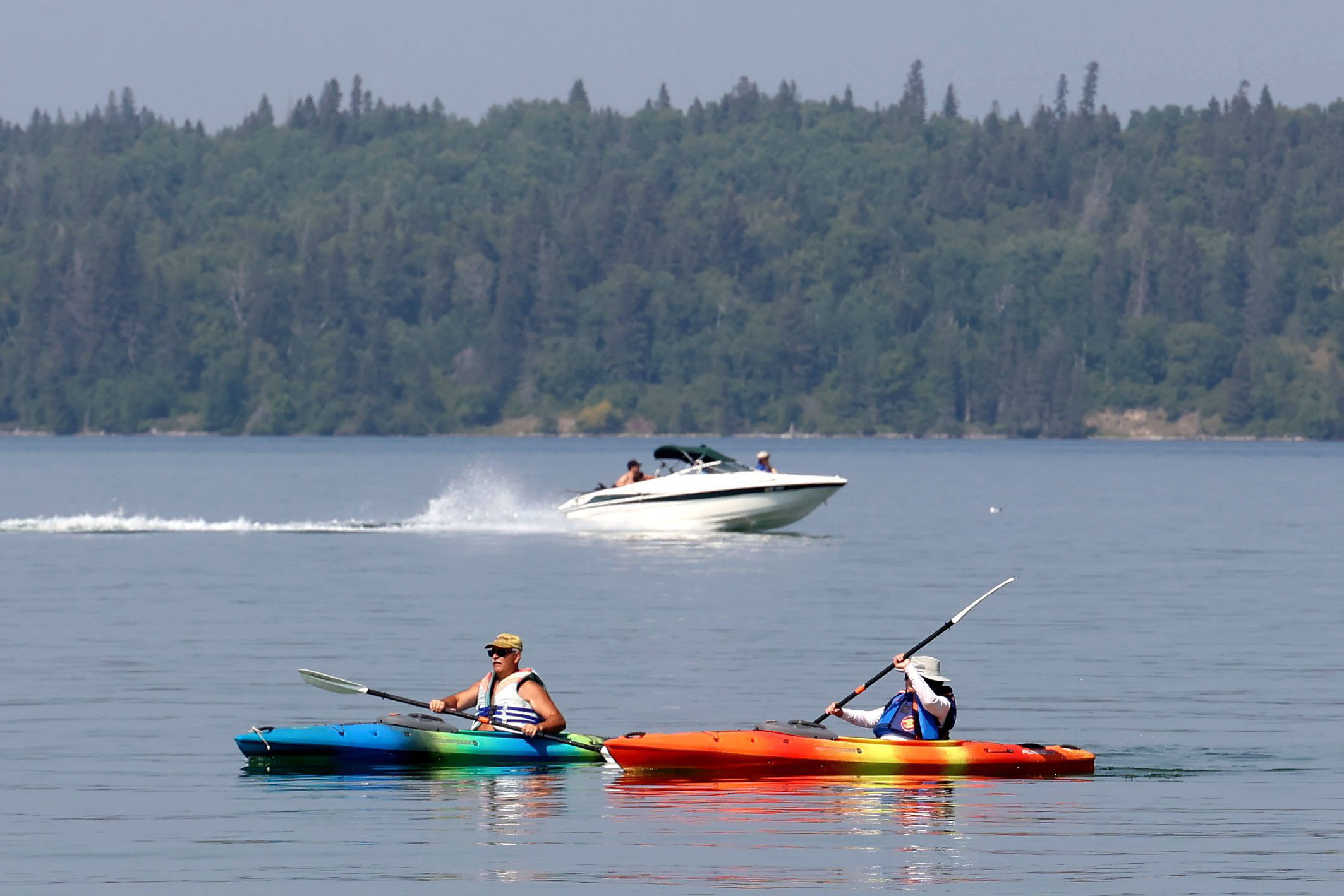 Kayakers paddle through Clear Lake while a boat passes behind them in Riding Mountain National Park, with a densely forested shore in the distance