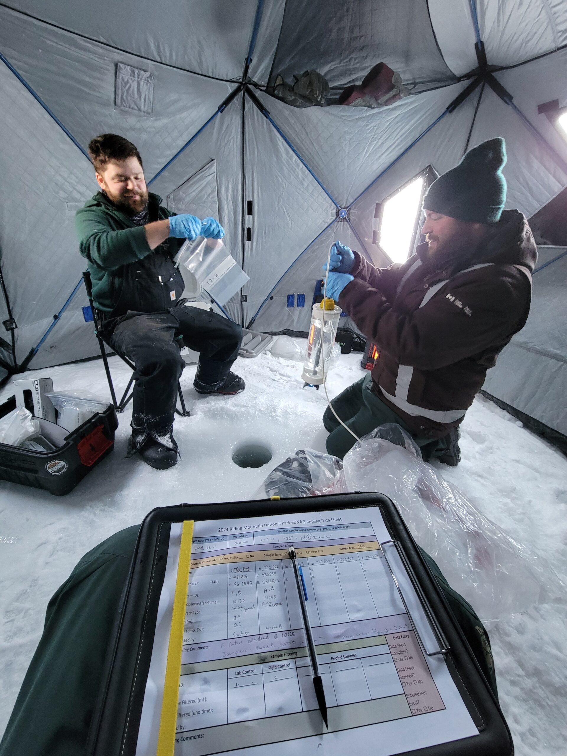 Riding Mountain National Park: Two Parks Canada staff members in uniform collect samples from a hole in the ice inside a tent structure over a frozen lake