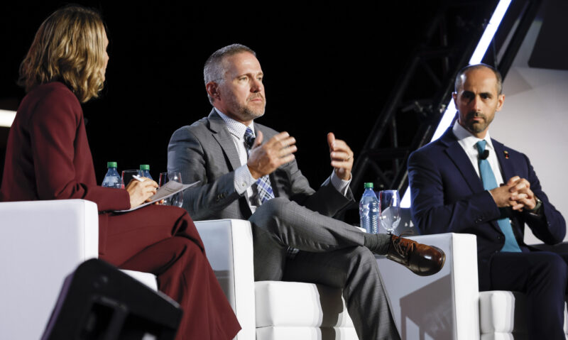 Pathways Alliance president Kendall Dilling, in a grey suit, speaks while sitting in a chair flanked by two other people on a stage.