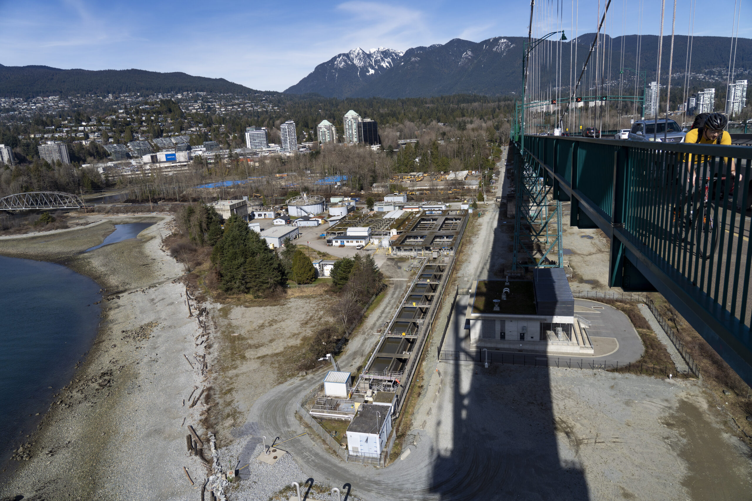A photo from above of the Lions Gate wastewater treatment plant, on the shores of Burrard Inlet. Cars and a cyclist are seen on the Lions Gate bridge is seen on the right side of the photo, with the North Shore mountains in the background
