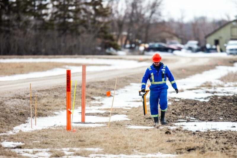 A man in high visibility blue coveralls and an orange hard hat walks along orange survey markers as he examines Imperial Oil's Winnipeg Products Pipeline in southern Manitoba